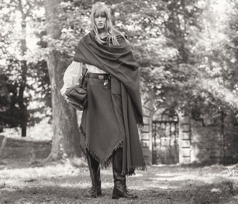 Monochrome portrait of a fashion model in vintage-style clothing, posing outdoors in a natural setting with a stone gate in the background.