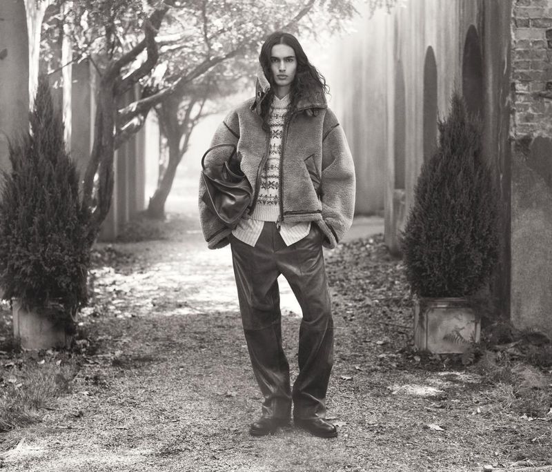Monochrome portrait of a male model in a shearling jacket and leather pants stands on a path amid greenery and classical architecture.