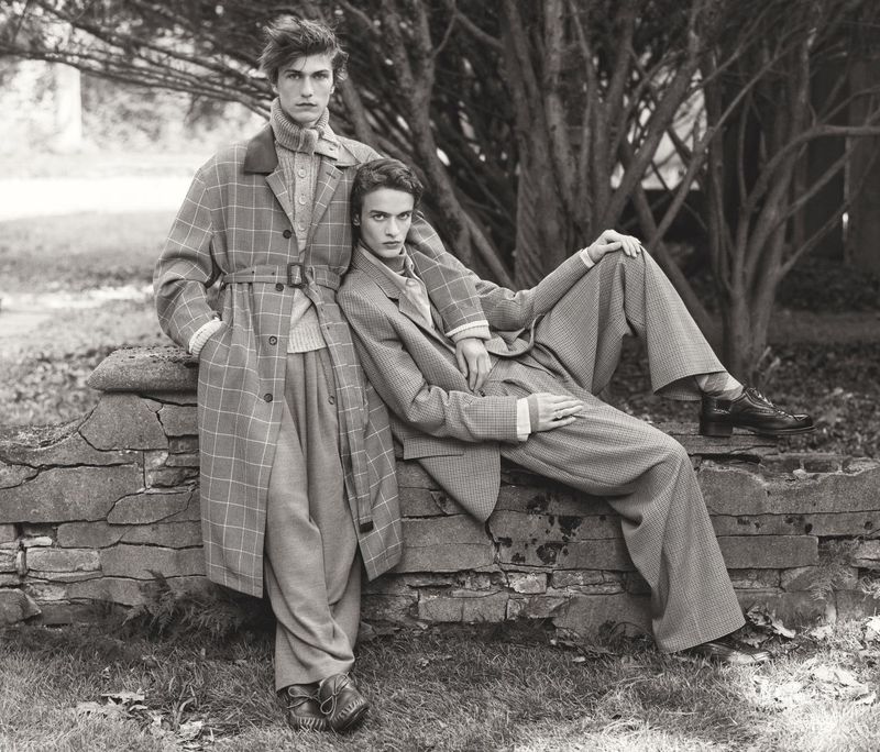 Two fashion models in monochrome outdoor shot. Man standing in trench coat, woman reclining in pantsuit against stone wall.