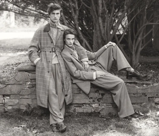 Two fashion models in monochrome outdoor shot. Man standing in trench coat, woman reclining in pantsuit against stone wall.