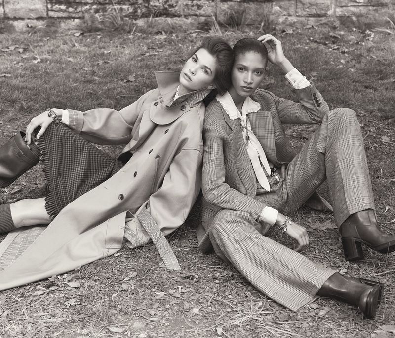 Two women in tailored outfits sit back-to-back in an outdoor setting with grass and a stone wall in the background in a fashion editorial.