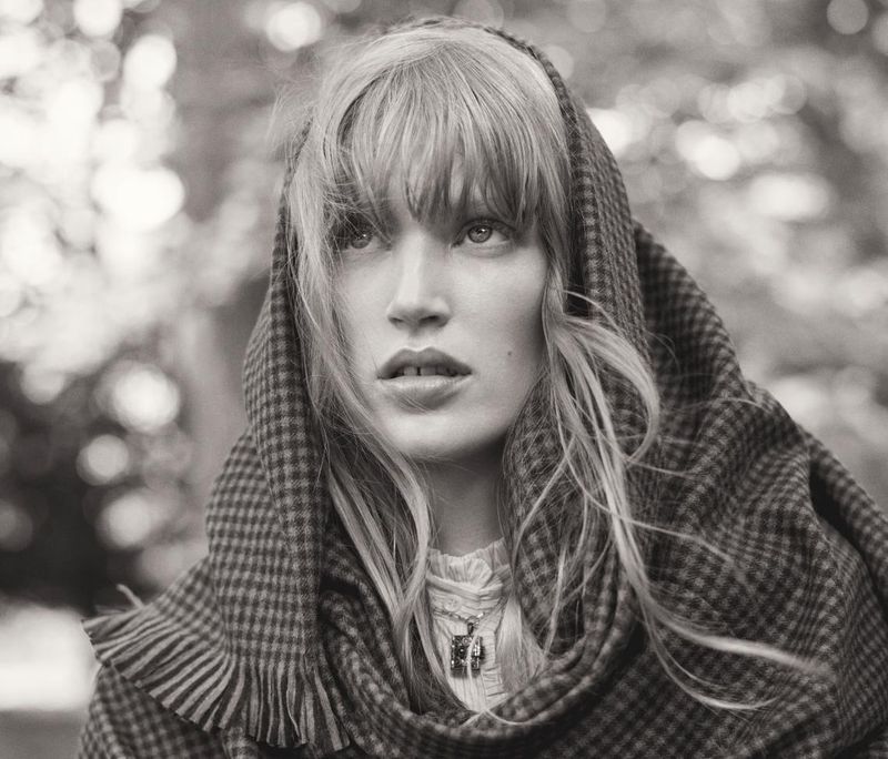Black-and-white portrait of a woman wearing a checkered fringed shawl draped over her head against a blurred outdoor background.