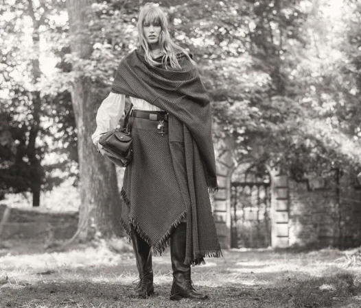 Model in patterned wrap shawl and tall boots holding a handbag outdoors by trees and a stone wall