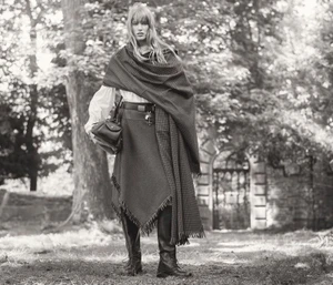 Model in patterned wrap shawl and tall boots holding a handbag outdoors by trees and a stone wall