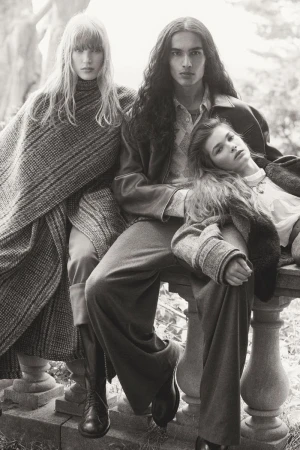 Black and white fashion campaign photograph of three models posed together by a stone balustrade outdoors.