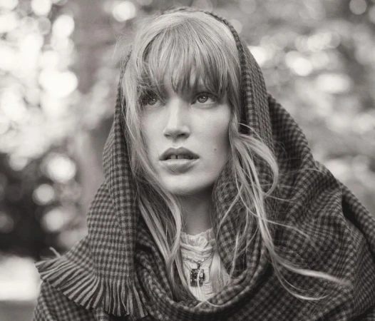 Black-and-white portrait of a woman wearing a checkered fringed shawl draped over her head against a blurred outdoor background.
