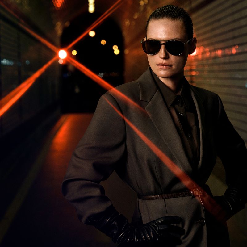 Woman in a grey suit and sunglasses poses in a tunnel illuminated by red laser lights, creating a dramatic and modern fashion statement.
