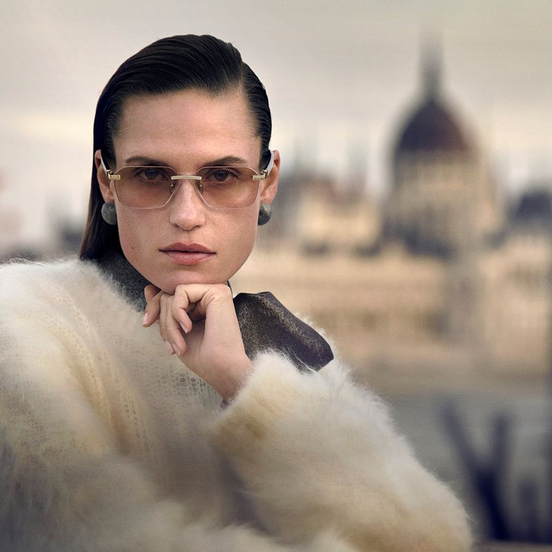 Model wearing tinted rimless glasses and a fluffy white sweater with chin resting on hand against an architectural urban backdrop.