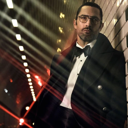Portrait of a man in a black tuxedo with glasses and a bow tie, leaning against a tiled wall in a tunnel with linear lights.