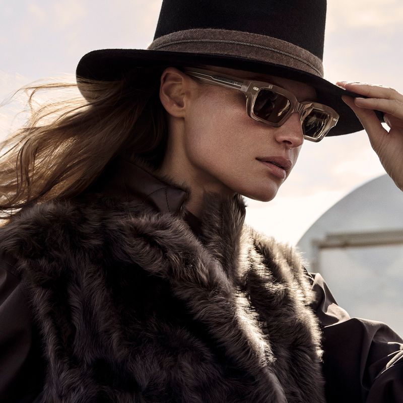 Model wearing clear rectangular sunglasses, a black wide-brim hat, and a fur vest looking to the side outdoors.