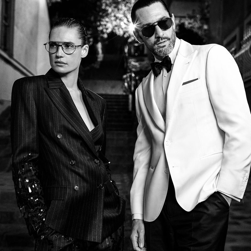 Black and white fashion shot of a woman in pinstripes and a man in a white tuxedo with bow tie, posing on an outdoor staircase.