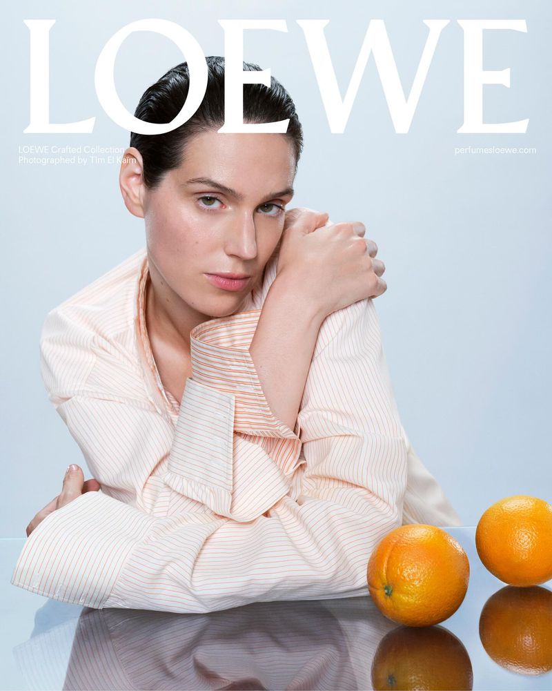 Elegant portrait of a woman with dark hair, wearing a striped shirt, two oranges reflecting on a glass table, Loewe brand signage overhead.