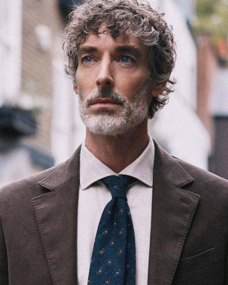 Portrait of a middle-aged man with grey hair, blue eyes wearing a brown jacket, white shirt and blue tie, posing outdoors.