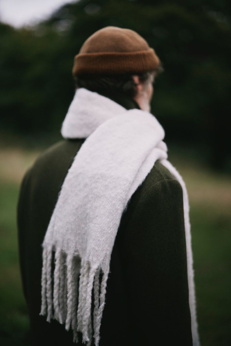 Back view of a person outdoors wearing a brown knit hat, olive green coat, and white textured scarf with fringe, in a soft, natural setting.