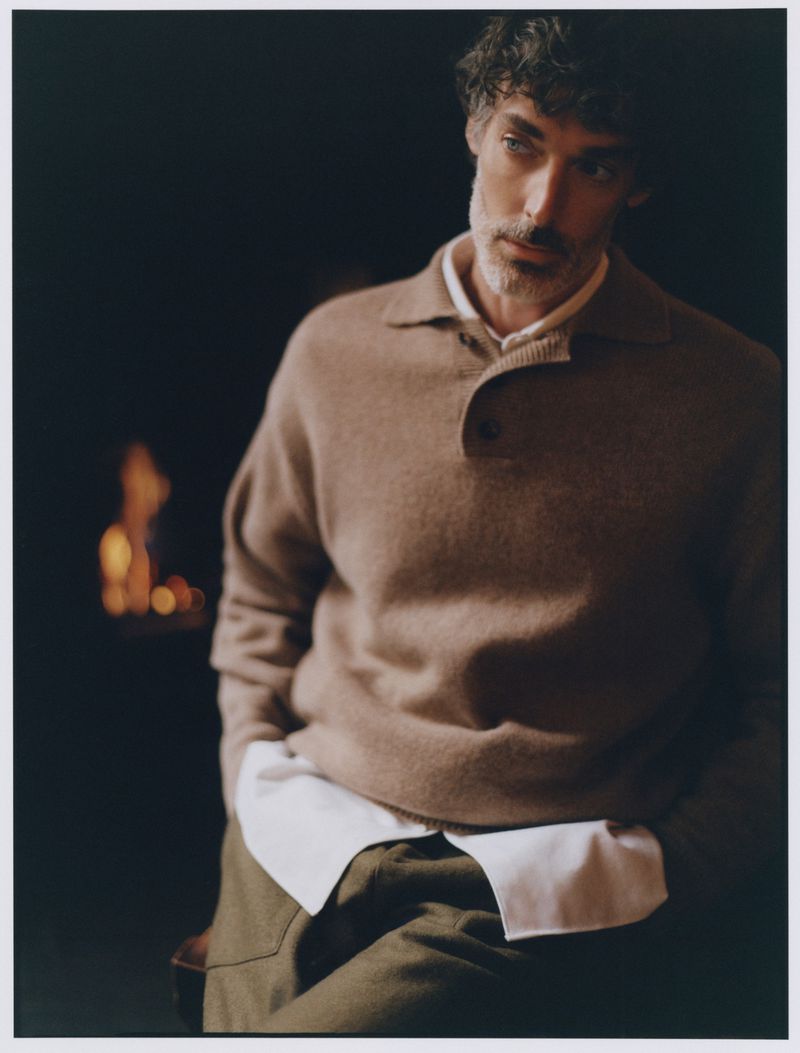 Portrait of a man in a beige collared sweater with a button, standing in front of a dark background with firelight.