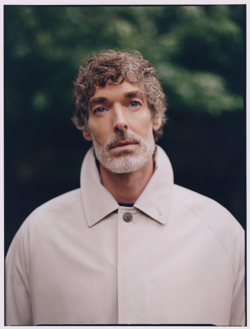 Portrait of a mature adult man with curly hair wearing a beige collared coat against a soft-focus green leafy background outdoors.