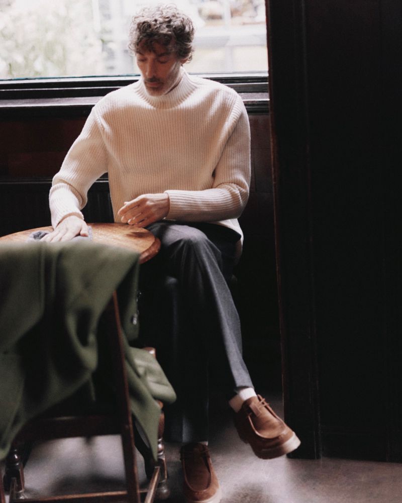 Man in a cream-colored turtleneck sweater sits by a window, wearing grey pants and brown shoes with a jacket draped on a chair.