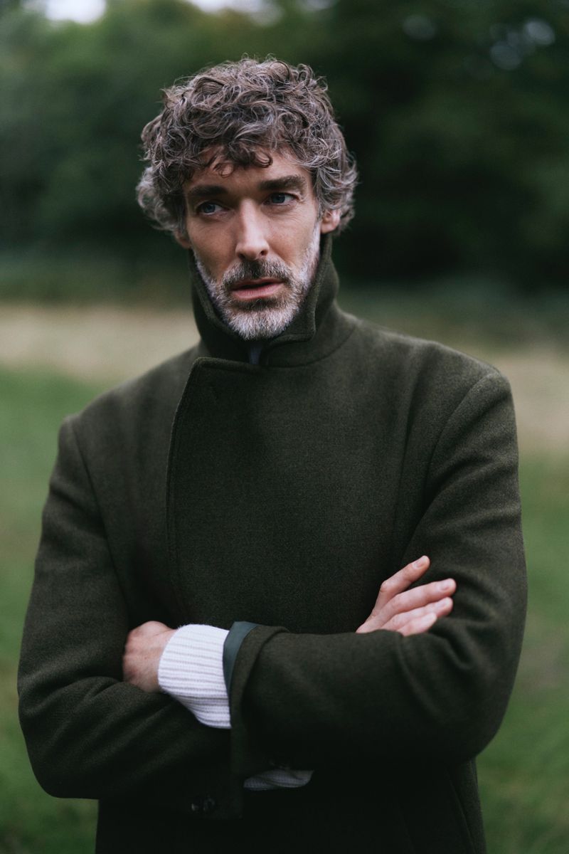 Portrait of a mature adult man with curly hair and a beard wearing a green coat with his arms crossed in an outdoor setting.