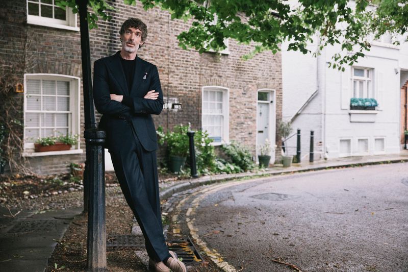 Man in a black suit leans against a pole on a city street, posing in front of brick buildings with green trees overhead and neutral light.