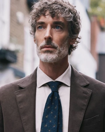 Portrait of model Richard Biedul wearing a brown suit jacket, white shirt, and blue patterned tie outdoors