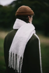 Man viewed from behind wearing a dark green coat, brown knit beanie, and thick textured off-white fringed scarf outdoors.
