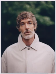 Model Richard Biedul wearing a high-collared off-white coat against a blurred green outdoor background.