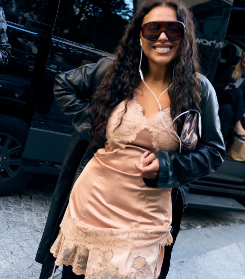 Smiling woman with long curly hair, wearing sunglasses, a slip dress with lace trim, and a black leather coat outside on a city street.