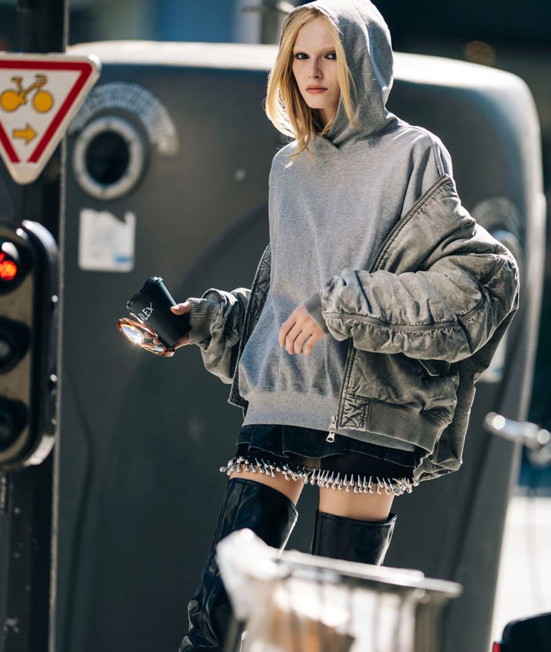 Alex Consani in a grey hooded sweatshirt and oversized grey bomber jacket with black patent boots on a city street.