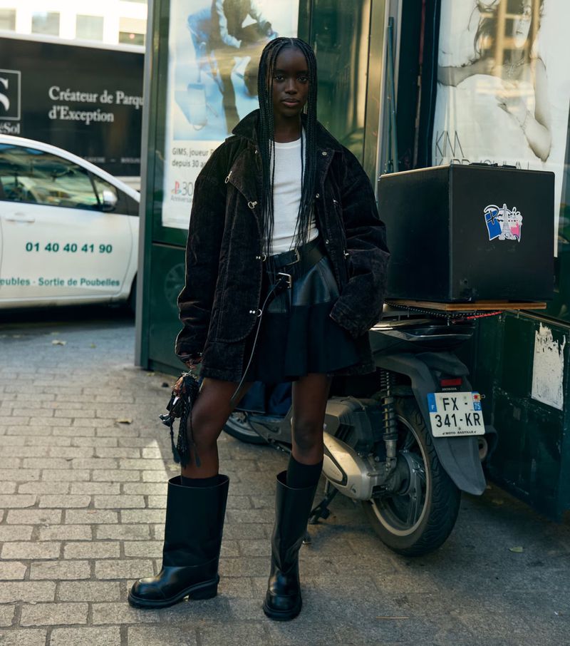A dark-skinned model in a white top, black skirt, jacket, and boots poses in an urban setting next to a moped. Fashion editorial.