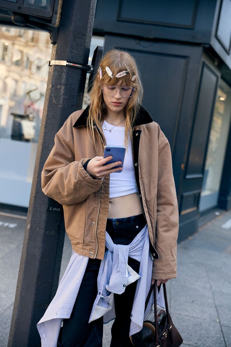 Street style featuring an adult woman with blonde hair, wearing a brown jacket, white top, and carrying a brown bag, looking at her phone.