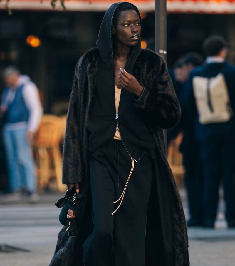 A stylish adult wears a faux fur coat over a hoodie and jogger pants, posing on an outdoor urban street, showcasing a modern fashion aesthetic.