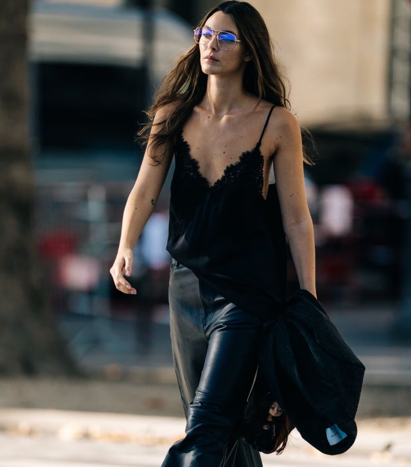 Adult woman in a black lace-trimmed top and shiny black pants walking in an outdoor urban setting, wearing blue-tinted glasses.
