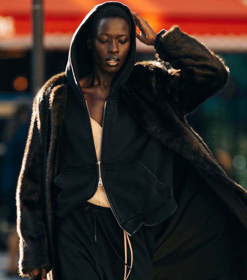 A stylish black woman wearing a fur coat and hooded jacket with dark pants poses outdoors with a neutral facial expression.