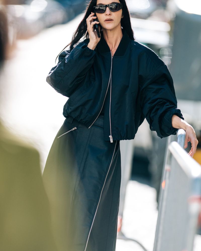 A stylish adult woman wearing a dark bomber jacket and long skirt talks on a cell phone outside on a sunny day.