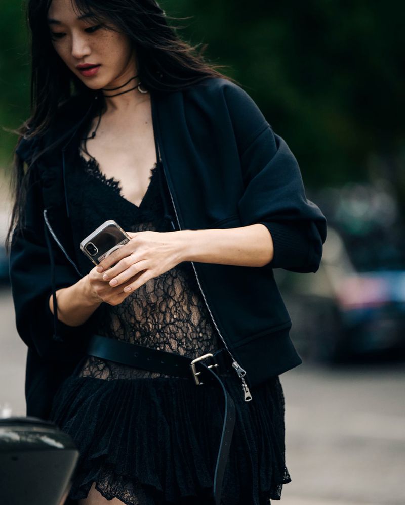 Asian woman in a black lace dress and bomber jacket looks at a phone outdoors in an urban setting with soft, diffused lighting.