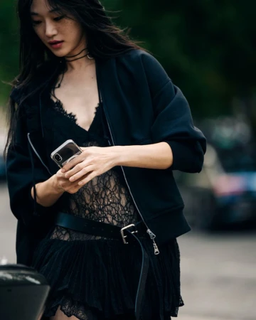 Asian woman in a black lace dress and bomber jacket looks at a phone outdoors in an urban setting with soft, diffused lighting.