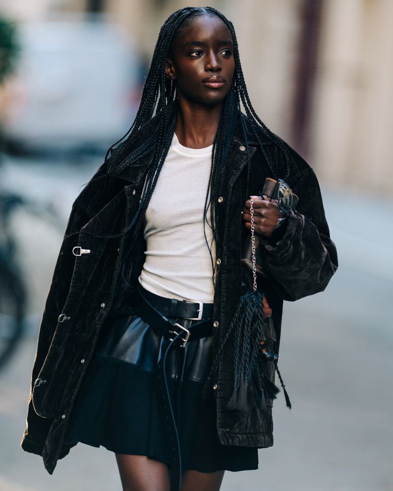 Fashionable Black woman in a jacket and skirt walking on the street. She carries a handbag with braids and wears stylish belts.