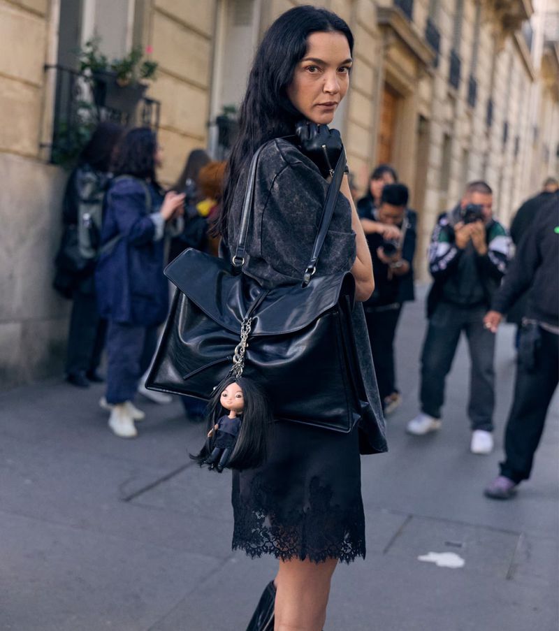 Woman with long dark hair and backpack with doll charm stands on a city street with photographers in the background on a cloudy day.