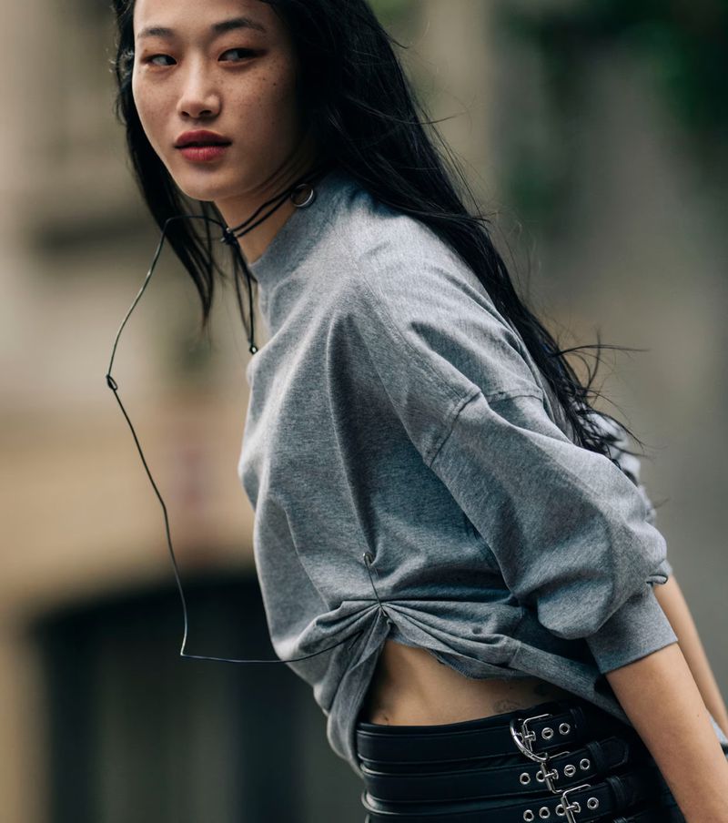 Portrait of a young Asian woman with long dark hair wearing a grey sweatshirt and black belt with silver buckles in a street setting.