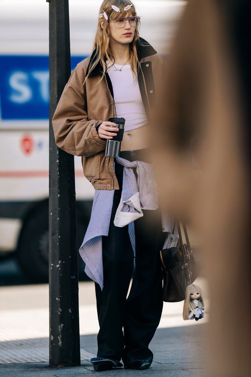 A slender woman with blond hair and glasses, wearing a brown jacket and black pants, stands on a city street holding a drink.