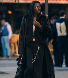Model Awar Odhiang walking on a city street wearing an oversized black fur coat over a black hooded jacket and track pants.