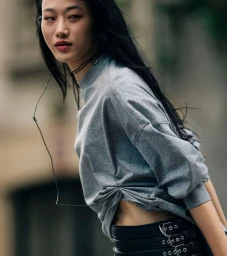 Model Sora Choi in a grey sweatshirt and multi-buckle black leather belt looking to the side outdoors.
