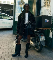 Black woman with braided hair wearing a dark coat and tall black boots standing on a cobblestone street