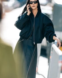 Model Mariacarla Boscono in black sunglasses and a dark bomber jacket talking on her phone outdoors for H&M In Betweens project.