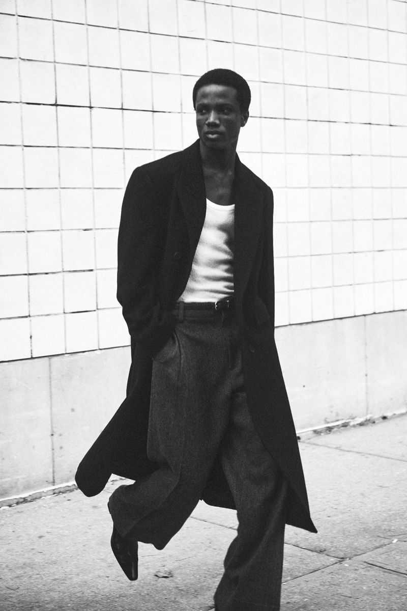 A black and white full shot of a Black male model walking outside wearing a long coat and wide-legged trousers, looking off to the side.