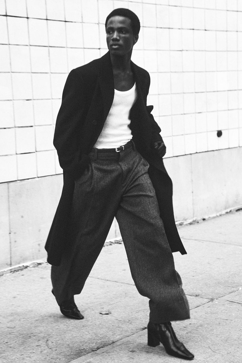 A Black man walks on a sidewalk in a black coat, white tank top, and grey slacks with a white tiled wall behind him in a fashion editorial.
