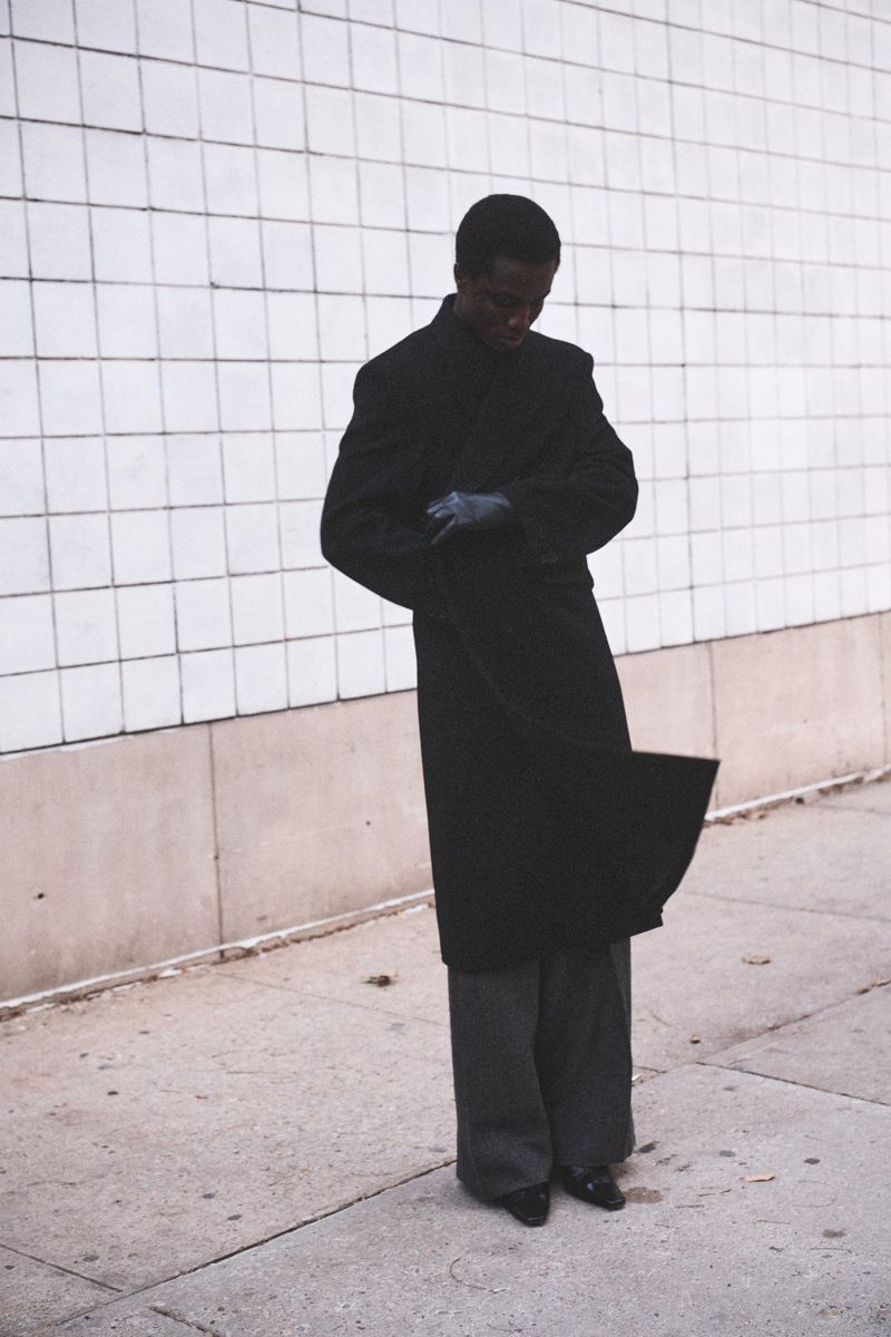 Model Mumin Jangani in a long black overcoat, wide-leg trousers, and leather gloves standing against a white tiled wall.