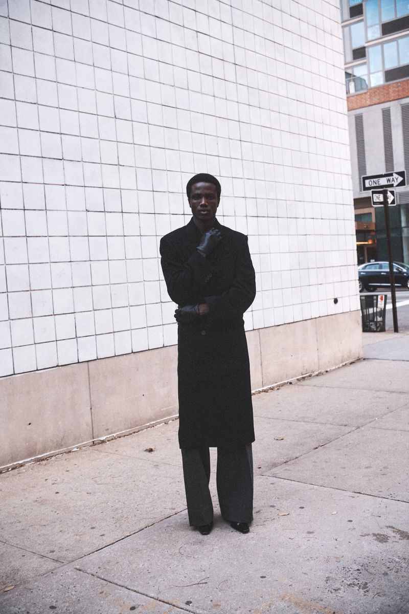 A dark-skinned adult male stands on a city sidewalk wearing a black coat and gloves, with a light grey tiled wall as a backdrop.