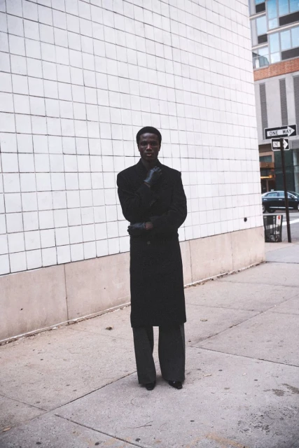 A dark-skinned adult male stands on a city sidewalk wearing a black coat and gloves, with a light grey tiled wall as a backdrop.