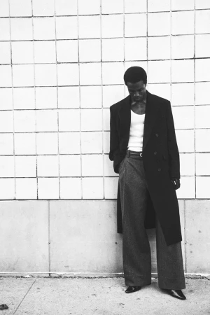 Model standing against a tiled wall wearing a long dark coat over a white tank top and grey trousers